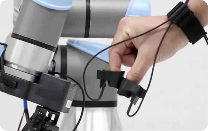 A hand wearing a black motion-tracking device interacts with a robotic arm, demonstrating a human-machine interface setup in a laboratory environment.
