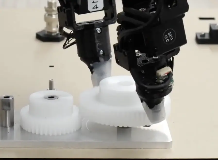 Close-up of a black robotic assembly tool positioning a white plastic gear on a metal fixture beside other gears in a precision workshop.