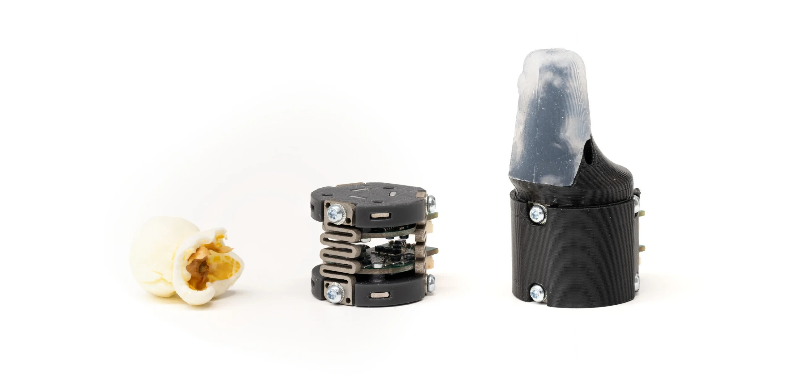Three objects on a white background: a popped kernel of popcorn, a small mechanical device, and a larger device with a translucent cover resembling a fingertip.